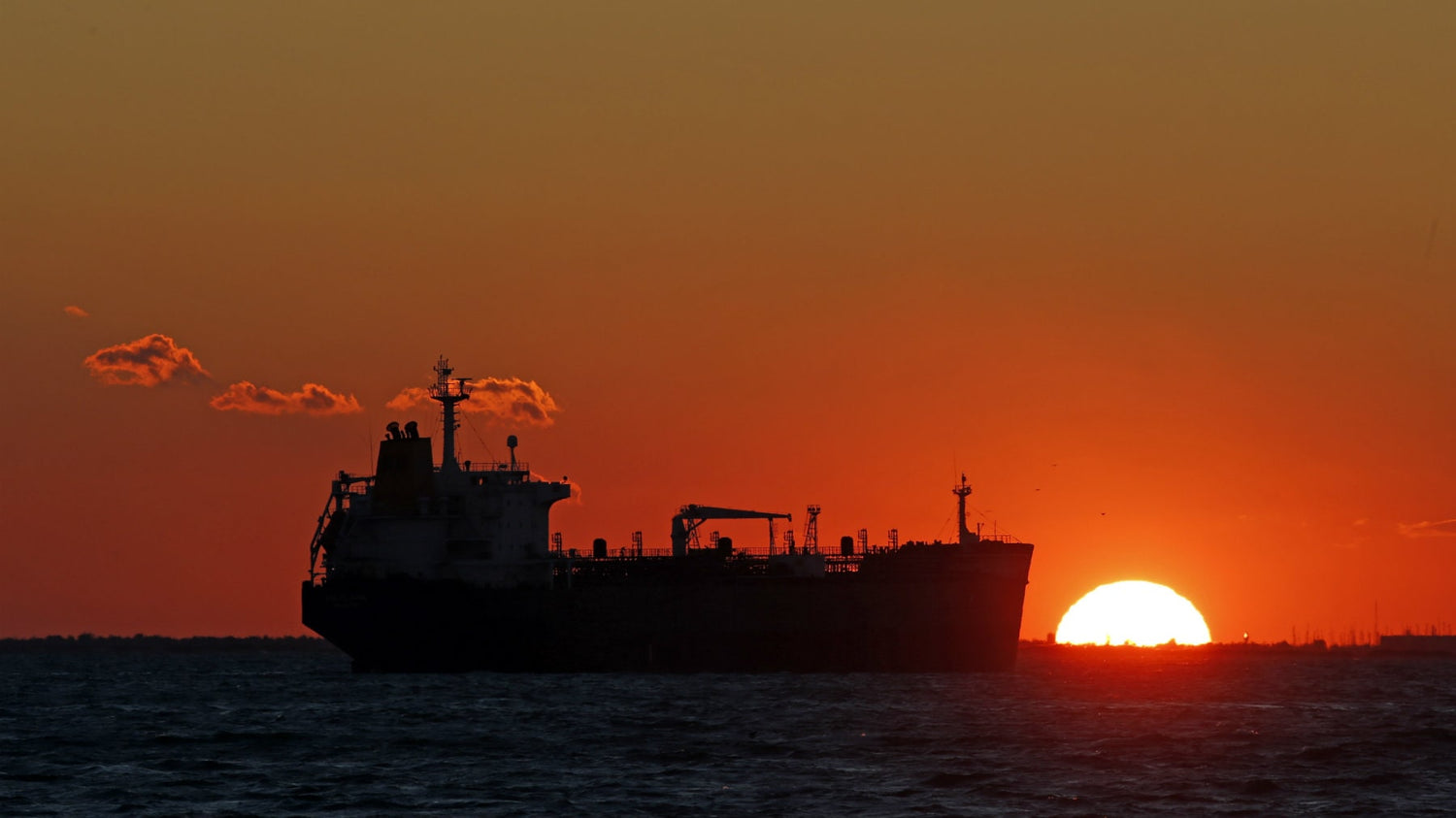 A vessel ship in the dark ocean besides a red setting sun and some clouds